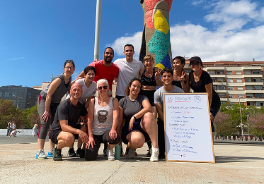 Grupo de personas sonriendo después de una sesión de entrenamiento al aire libre en Barcelona, con una pizarra que muestra la rutina de ejercicios y una escultura colorida al fondo.